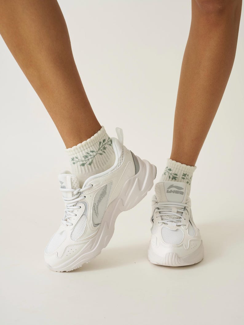 May include: White trainers with a chunky sole and mesh detailing. The shoes are paired with white socks featuring a green floral pattern. The trainers have the brand name "Li-Ning" on the tongue.