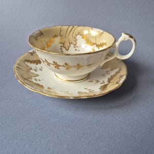Può includere: Un set di tazza da tè e piattino in porcellana color crema con un delicato motivo a foglie dorate. La tazza presenta un bordo smerlato, un manico curvo e una base rialzata. Il piattino è abbinato alla tazza con bordo smerlato e motivi floreali.