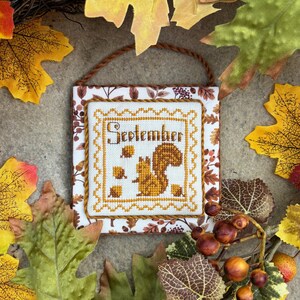 May include: A cross-stitched wall hanging with the word "September" and a squirrel design. The artwork is framed with a decorative border and a brown cord for hanging. Autumn leaves and berries surround the artwork.