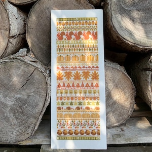 May include: A white cross-stitch pattern with an autumnal theme. The pattern features a variety of autumnal elements, including pumpkins, leaves, squirrels, and acorns. The pattern is framed by a border of brown and yellow.