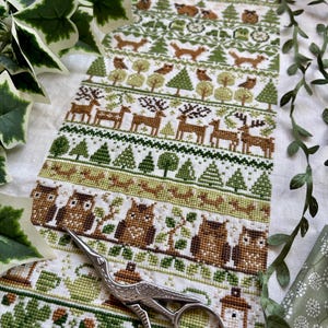 Puede incluir: Un detallado patrón de punto de cruz con animales del bosque y árboles en tonos verdes y marrones. El diseño incluye ciervos, zorros, búhos y varios patrones de árboles. Un par de tijeras plateadas descansa sobre la tela.