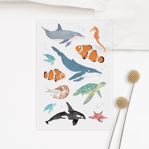 Könnte beinhalten: Ein Set mit bunten Meerestier-Stickern, darunter ein Blauwal, ein Delfin, ein Seepferdchen, eine Schildkröte, ein Nautilus, eine Orca und zwei Clownfische.