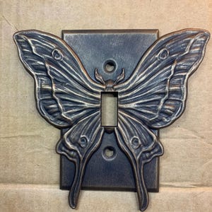May include: A decorative light switch cover featuring a butterfly design. The cover is made of metal with a dark bronze finish. The butterfly wings extend outward from the rectangular base, with detailed wing patterns. The switch opening is centered.