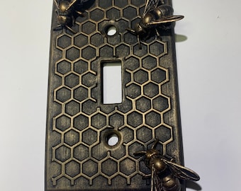 May include: A rectangular light switch cover plate featuring a raised honeycomb pattern in a dark bronze finish. Three dimensional bee figures are attached to the surface, with two bees at the top corners and one bee at the bottom right.