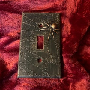 Könnte beinhalten: Dekorative Lichtschalterabdeckung mit Spinnen- und Spinnennetz-Design. Die rechteckige, dunkelgraue Abdeckung hat ein strukturiertes Netzmuster. Eine bronzefarbene Spinne ist angebracht. Die Abdeckung hat eine einzelne Schalteröffnung und zwei Schraubenlöcher.