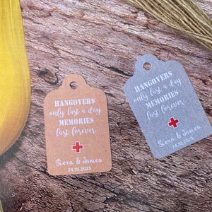May include: Two wedding favor tags with the text "Hangovers only last a day Memories last forever" and a red plus sign. The tags are on a wooden surface. The tag on the left is brown and the tag on the right is silver.