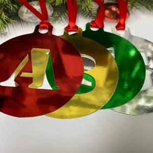 Peut inclure: Quatre décorations de Noël avec les lettres A, B, C et D découpées. Les décorations sont rouge, or, vert et argent.