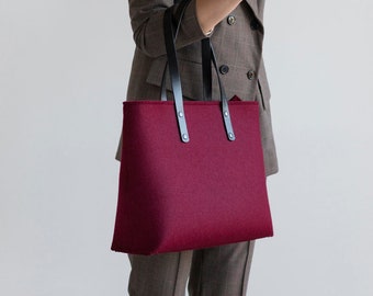 May include: A burgundy felt tote bag with black leather handles and a top zipper closure. The bag is being held by a person wearing a brown blazer.