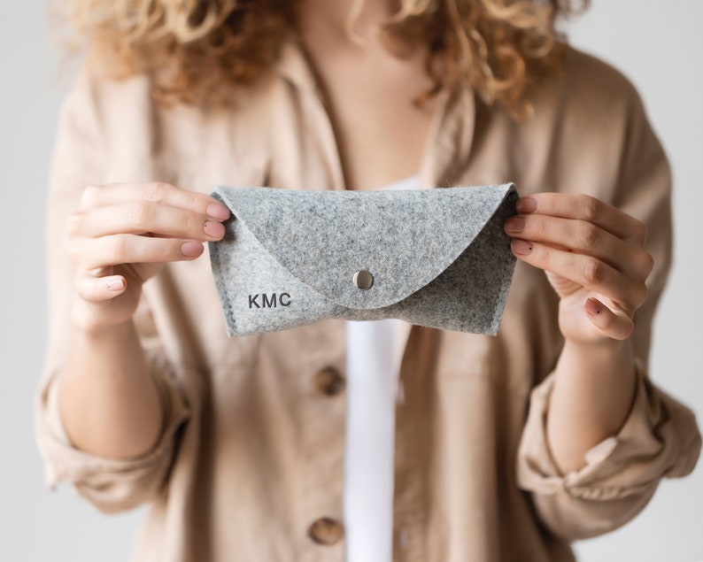 May include: A gray felt glasses case with a silver button closure and the initials "KMC" embroidered on the front.