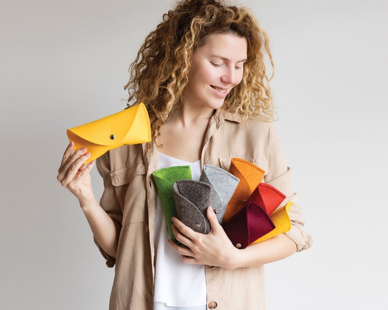 May include: A woman holds a variety of felt pouches in different colors, including yellow, green, gray, brown, orange, red, and burgundy. The pouches are folded in a fan shape.