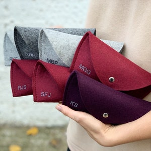 May include: Six felt pouches in various shades of gray and red with personalized initials embroidered on the front. The pouches are folded in half and held in a hand.