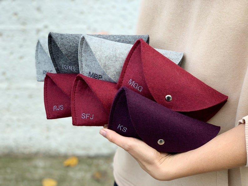 May include: Six felt pouches in various shades of gray and red with personalized initials embroidered on the front. The pouches are folded in half and held in a hand.