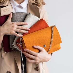 Puede incluir: Un conjunto de cuatro bolsos de fieltro en varios colores, incluyendo rojo, gris, beige, naranja y una correa de cadena plateada.