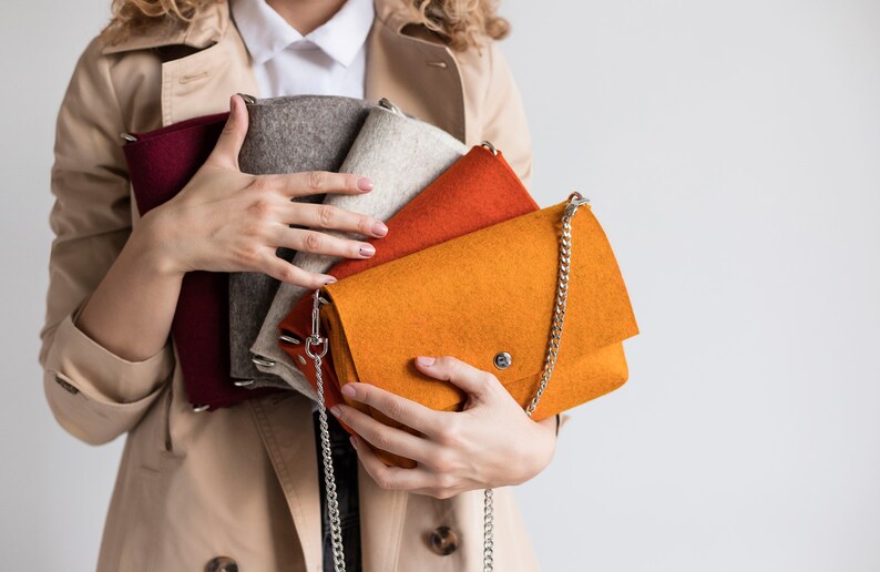Puede incluir: Un conjunto de cuatro bolsos de fieltro en varios colores, incluyendo rojo, gris, beige, naranja y una correa de cadena plateada.
