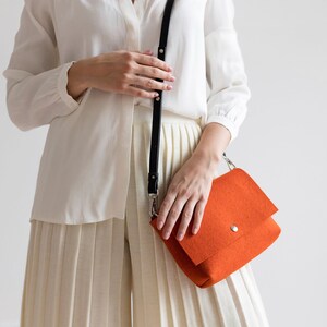 Puede incluir: Una mujer con una camisa blanca y una falda blanca plisada sostiene un bolso bandolera de color naranja brillante con una correa negra.