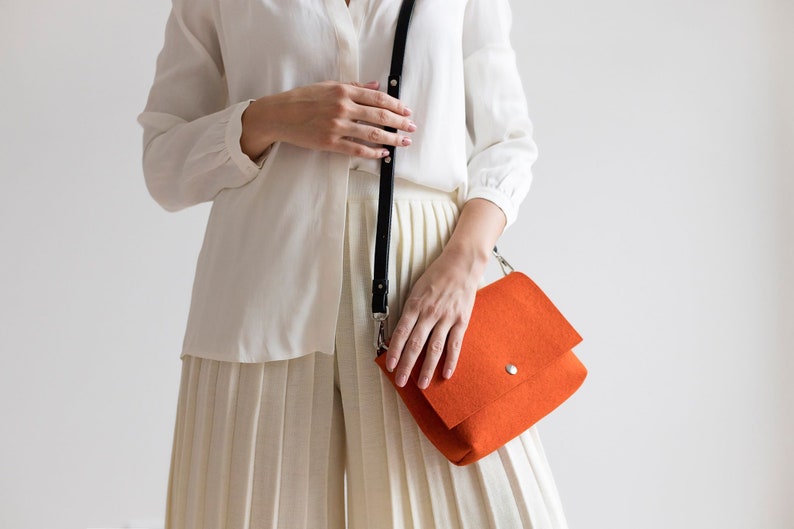 Puede incluir: Una mujer con una camisa blanca y una falda blanca plisada sostiene un bolso bandolera de color naranja brillante con una correa negra.