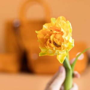 Puede incluir: Un solo tulip&aacute;n amarillo con p&eacute;talos rizados, la flor se sostiene en una mano sobre un fondo naranja borroso.