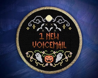 May include: A black and white cross-stitch embroidery hoop with a Halloween theme. The hoop features a black background with white stitching of a moon, bats, ghosts, and a pumpkin. The text "1 New Voicemail" is stitched in white on a black background.