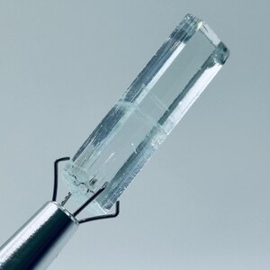 Könnte beinhalten: Ein klarer, rechteckiger Kristall, der von einem silbernen Metallwerkzeug gehalten wird. Der Kristall hat einen hellblauen Farbton und erscheint transparent mit sichtbaren inneren Strukturen. Das Werkzeug hat einen zylindrischen Griff und Drahtzinken.
