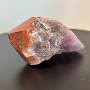 May include: A natural crystal formation with a striking colour contrast. The faceted top is a rich orange-red, blending into a deep purple base. The crystal has a rough, textured surface, highlighting its natural form. A unique mineral specimen.