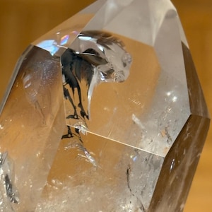 Op de afbeelding: Close-up van een heldere bergkristal punt met interne insluitsels. De kristal heeft een facetvormige, geometrische vorm met een scherpe apex. Het interieur heeft donkere, organisch ogende patronen. De kristal staat tegen een warme, neutrale achtergrond.