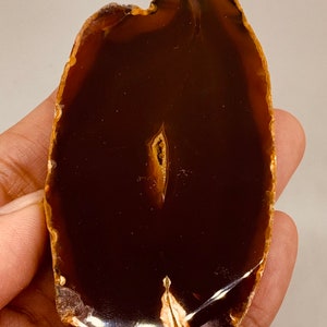 Könnte beinhalten: Eine braune Agatscheibe mit einer natürlichen, unregelmäßigen Form und einer durchscheinenden, glasartigen Oberfläche. Der Achat hat ein charakteristisches, wirbelndes Muster aus braunen und weißen Bändern.