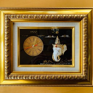 Könnte beinhalten: Eine goldgerahmte Wanduhr mit einer weißen Elefantenfigur und einem Zifferblatt mit römischen Ziffern. Die Uhr ist vor einem schwarzen Hintergrund mit goldener Umrandung platziert.