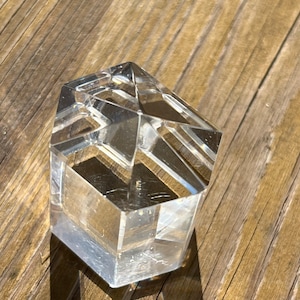 Peut inclure: Un prisme hexagonal en verre transparent avec un sommet pointu. Le prisme est posé sur une surface en bois.