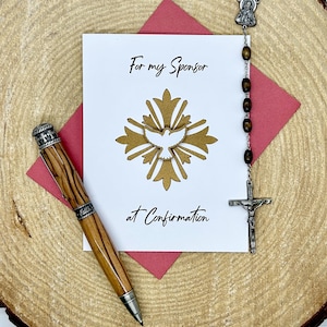 May include: A Confirmation card with the text "For my Sponsor at Confirmation" and a gold dove design. A wooden pen and a rosary with a crucifix are also present, all arranged on a wooden surface.