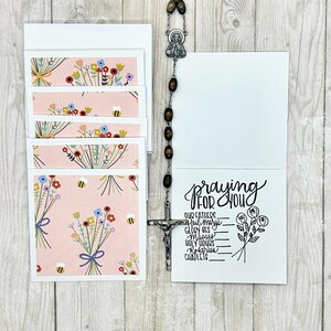 May include: A set of blank greeting cards with a floral design and a rosary. The cards feature a pink background with illustrations of flowers and bees. One card is open, displaying the words "Praying for You" and a list.