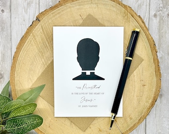 May include: A white card with a black silhouette of a priest's head and collar, with the text "THE Priesthood IS THE LOVE OF THE HEART OF Jesus. ST. JOHN VIANNEY." A black and gold pen rests on a wooden slice.