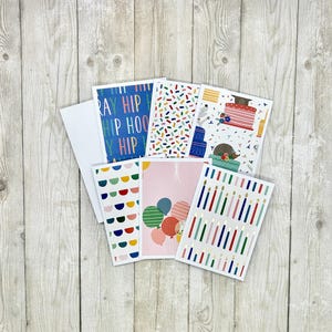May include: A collection of six rectangular greeting cards featuring various celebratory designs including the text HIP HIP HOORAY, colorful sprinkles, birthday cakes, semi-circles, balloons, and vertical candles in shades of blue, red, and green.
