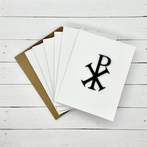 May include: A stack of white greeting cards with a black Chi Rho symbol on the front, fanned out over a brown envelope. The cards are on a white wooden surface.