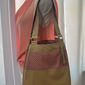 May include: A beige tote bag with a red and white checkered pattern and a beige bottom. The bag has beige straps and a decorative tassel. The bag is displayed on a mannequin.
