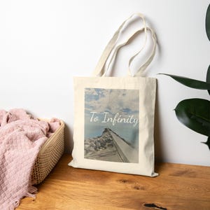 May include: A beige tote bag with a scenic image of a stone pier extending into the ocean under a cloudy sky. The words "To Infinity" are written across the image in a cursive font. The bag has long handles.