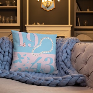 May include: A blue and pink throw pillow with the numbers 1943 and a pearl necklace design. There are also pink butterflies on the pillow.