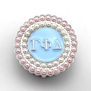 Puede incluir: Un pin redondo plateado con un centro azul claro y las letras "ΓΦΔ" en blanco. El pin está rodeado por un anillo de perlas rosas y blancas.
