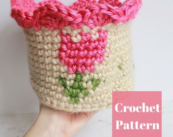 May include: Crochet pattern for a cream-colored basket with pink and green flowers. The basket has a scalloped pink edge.