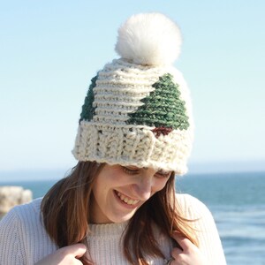 May include: A white crocheted beanie with a white pom pom on top. The beanie has a green and brown crocheted design of a Christmas tree on the front.