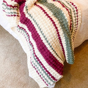May include: A striped crocheted throw blanket in shades of cream, burgundy, and gray. The blanket is folded and draped over a white bed.