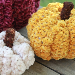 May include: Three crocheted pumpkins in different colors: burgundy, yellow, and white. The pumpkins are made with a fuzzy yarn and have a textured surface.