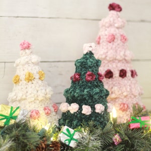 Puede incluir: Tres &aacute;rboles de Navidad de crochet decorados con rosetas de terciopelo rosa, blanco y verde. Los &aacute;rboles est&aacute;n dispuestos sobre una superficie de madera blanca con una guirnalda verde y peque&ntilde;os regalos envueltos.