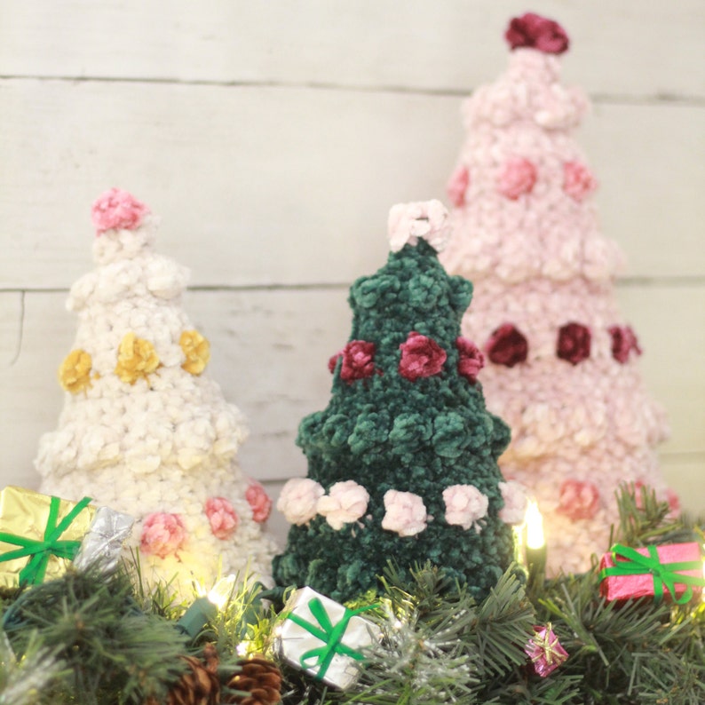 Puede incluir: Tres &aacute;rboles de Navidad de crochet decorados con rosetas de terciopelo rosa, blanco y verde. Los &aacute;rboles est&aacute;n dispuestos sobre una superficie de madera blanca con una guirnalda verde y peque&ntilde;os regalos envueltos.