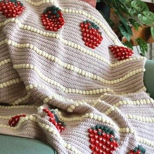 K&ouml;nnte beinhalten: Eine rosa-wei&szlig;e H&auml;keldecke mit Erdbeermuster. Die Decke liegt auf einem t&uuml;rkisfarbenen Sofa.