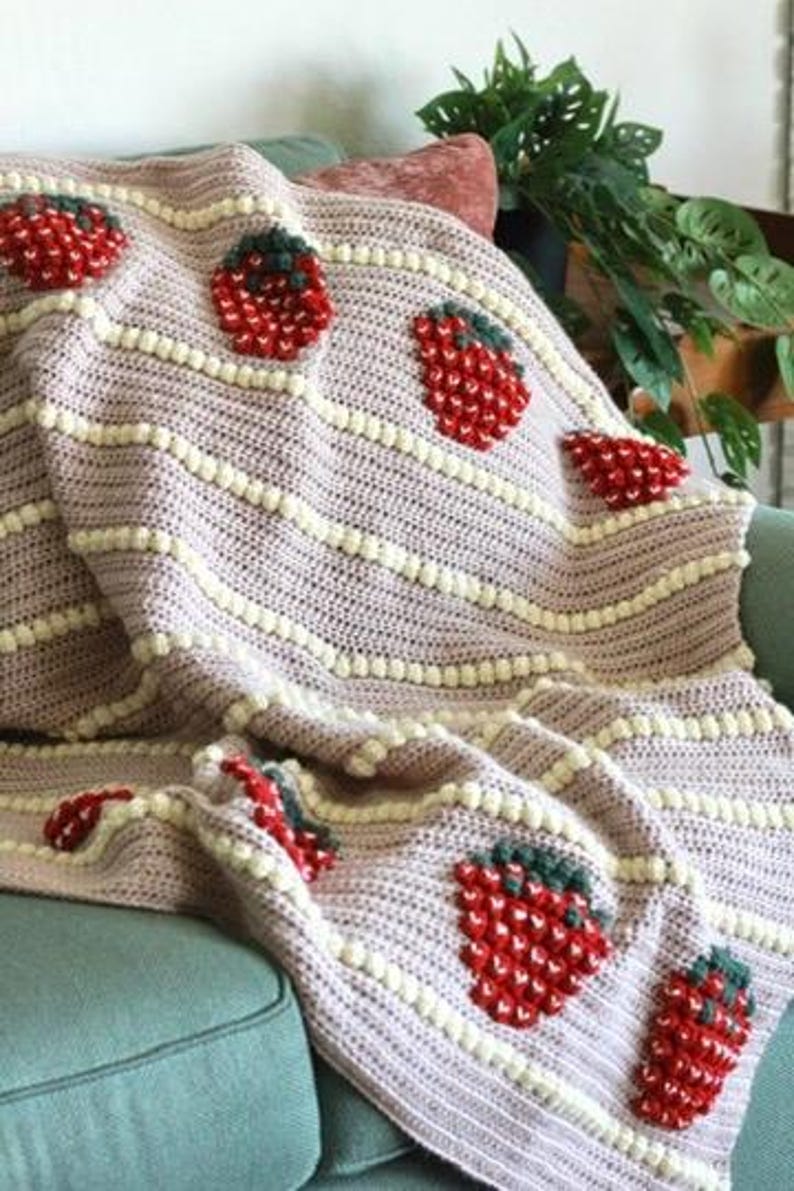 K&ouml;nnte beinhalten: Eine rosa-wei&szlig;e H&auml;keldecke mit Erdbeermuster. Die Decke liegt auf einem t&uuml;rkisfarbenen Sofa.