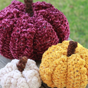 May include: Three crocheted pumpkins in different sizes and colors. The largest pumpkin is a deep burgundy color, the medium pumpkin is a bright yellow, and the smallest pumpkin is a light cream color.