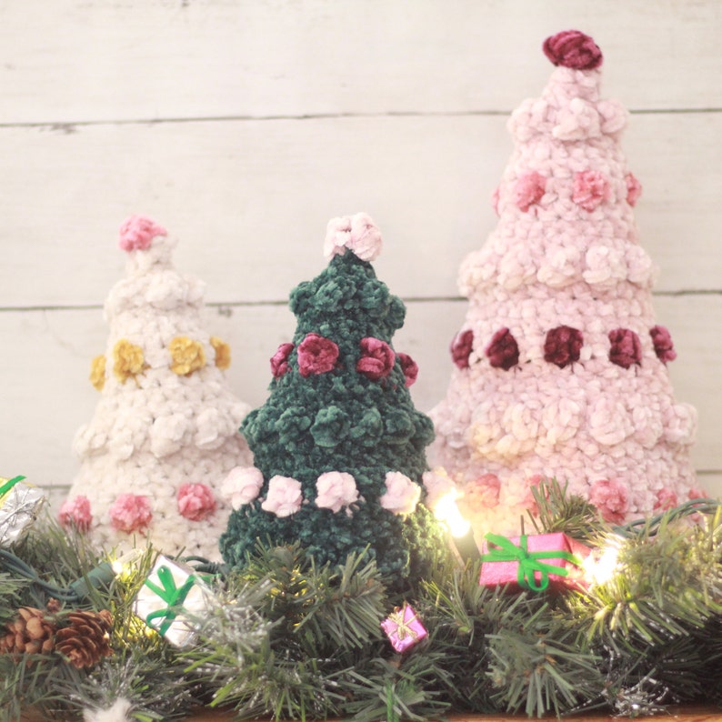 Puede incluir: Tres &aacute;rboles de Navidad de crochet decorados con rosetas de terciopelo rosa, verde y burdeos. Los &aacute;rboles est&aacute;n rodeados de una guirnalda de ramas de pino y luces blancas.