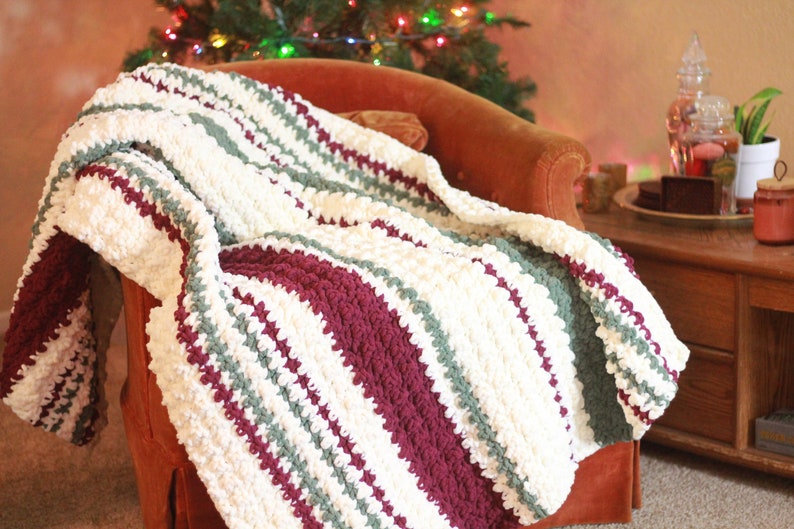 May include: A white, green, and burgundy striped crocheted throw blanket draped over a red armchair. The blanket has a textured, puffy look.