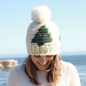 May include: A white knit beanie with a green and brown tree design and a white pom pom on top.