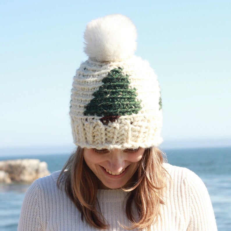 May include: A white knit beanie with a green and brown tree design and a white pom pom on top.